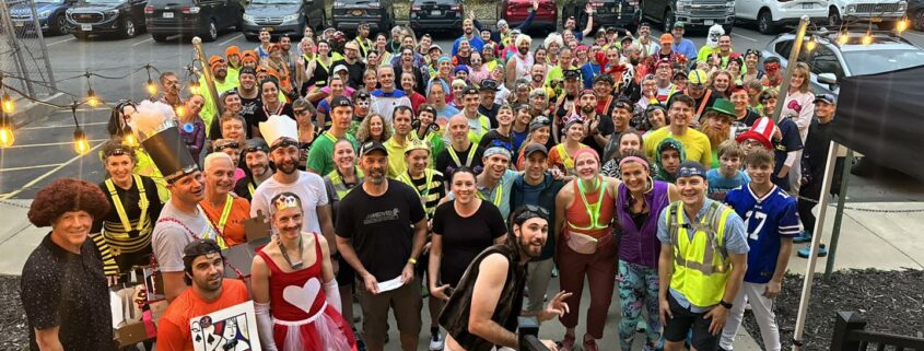 A large, diverse group of participants, many wearing Halloween costumes, pose for a photo at a running event. Costumes visible in the front include a man in a bee costume, a woman dressed as a Queen of Hearts, and a person holding a large cutout of a playing card.