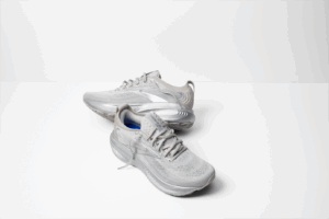 A pair of silver and white Brooks Adrenaline 25 running shoes are displayed against a plain white background. The shoe in the foreground is seen from a slightly elevated side angle, with its laces neatly tied. The second shoe is positioned behind it, also visible from a similar side angle, but a bit more elevated and angled towards the back. The lighting is even, highlighting the sleek design and metallic accents of the shoes.