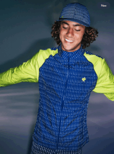 A person with curly brown hair and a tan complexion smiles while wearing a blue and neon yellow long-sleeved athletic jacket and a matching blue baseball cap. The jacket and cap have a pattern of thin white vertical lines. The background is a gradient of dark blue and purple.