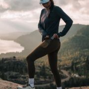 A person in athletic wear, including leggings and a long-sleeved shirt, reaching for something in their back pocket while standing on a rocky outcrop overlooking a lake and mountains.