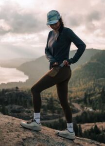 A person in athletic wear, including leggings and a long-sleeved shirt, reaching for something in their back pocket while standing on a rocky outcrop overlooking a lake and mountains.
