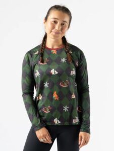 A smiling woman with braided pigtails wears a long-sleeved green and black argyle pattern shirt, dotted with snowflakes and various festive images of bunny rabbits in holiday attire.