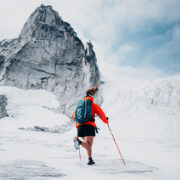 A person wearing an orange jacket, black shorts, and a gray backpack is seen from behind, walking across a vast, snow-covered landscape with a towering, jagged mountain in the background. They are using red trekking poles for support.