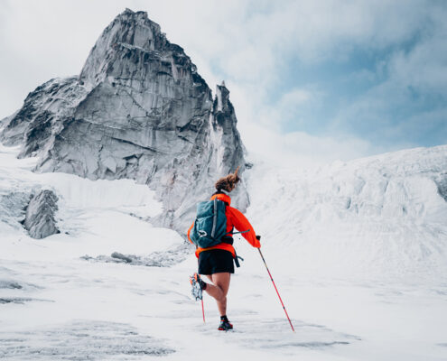 A person wearing an orange jacket, black shorts, and a gray backpack is seen from behind, walking across a vast, snow-covered landscape with a towering, jagged mountain in the background. They are using red trekking poles for support.